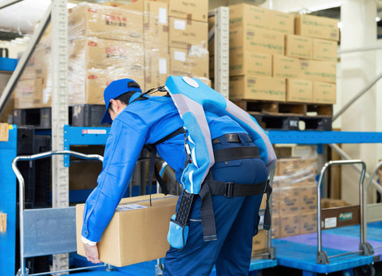 Ein Arbeiter in blauer Uniform hebt mit einem tragbaren Exoskelett einen Karton in einem mit Kartons gefüllten Lagerhaus. - © Foto: metamorworks - stock.adobe.com